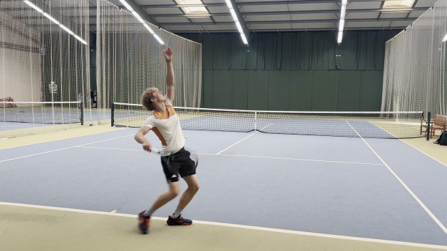 An indoor court filmed from behind the baseline at an angle. The player is serving with their back half to the camera.